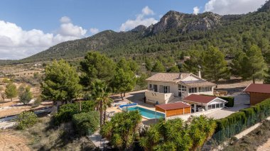 Engebeli dağların ve orman manzarasının yakınındaki havuzu ve ağaçları olan zarif bir villa.