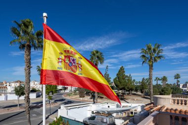 Palmiye ağaçlarının üzerinde dalgalanan İspanyol bayrağı açık mavi gökyüzünün altında sokak ve evleri