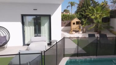 Yüzme havuzlu modern bahçe, mobilyalar ve palmiye ağaçlarıyla çevrili ahşap çocuk oyun evi.