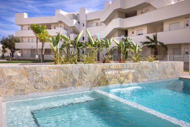 Şelale özellikli modern havuz, tropikal bitkiler ve arka planda beyaz apartmanlar