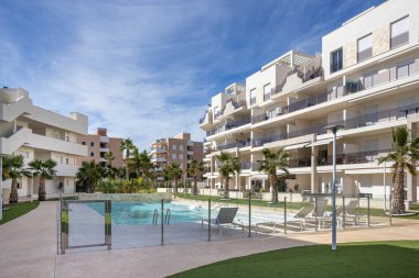 Cam çitli havuzu ve palmiye ağaçlarıyla çevrili şezlongları olan güneşli bir apartman.