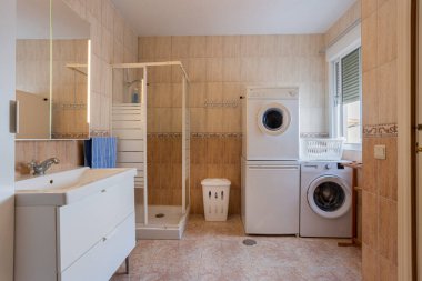 Duş, çamaşır makinesi, kurutma makinesi, lavabo dolabı ve doğal pencere ışığı olan geniş bir banyo.