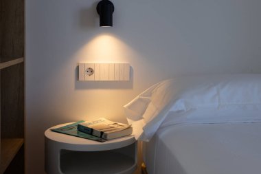 Kitaplı minimalist komodin, duvar ışığı, beyaz yatak ve modern sıcak ortam.