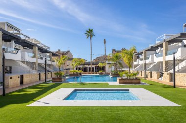 İki modern havuz, güneşli gökyüzünün altında palmiye ağaçları ve çağdaş apartmanlar ile çevrili..