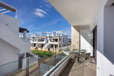 Geniş balkon, cam parmaklıklar, masa ve sandalyeler, modern beyaz konut kompleksi.