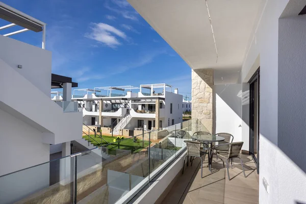 Geniş balkon, cam parmaklıklar, masa ve sandalyeler, modern beyaz konut kompleksi.