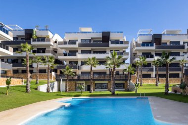 Havuzu, palmiye ağaçları, balkonları ve mavi gökyüzünün altında yeşil çimenlikleri olan çağdaş apartman kompleksi.