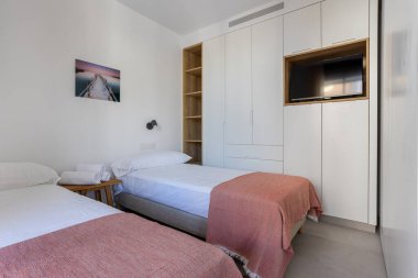 Parlak yatak odası, çift kişilik yataklar, beyaz gömme gardırop, TV ünitesi, ve basit rahat dekor.