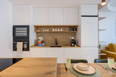 Parlak beyaz mutfak, ahşap kaplama, siyah musluk, kahve istasyonu ve dahili fırın.