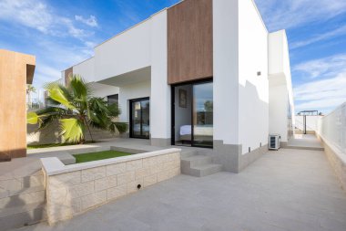 Geniş pencereli modern ev cephesi, palmiye ağacı peyzajı, fayanslı veranda ve temiz çizgiler.