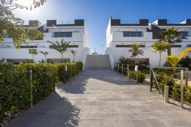 Modern konut gelişiminde peyzaj yolu, merkez merdiven ve minimalist beyaz cepheli çağdaş konut binalarının parlak güneşli manzarası.