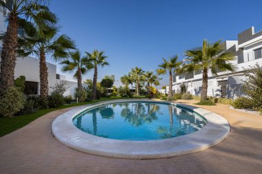 Çağdaş beyaz apartmanlar, açık mavi gökyüzünün altında, palmiye ağaçları ve manzaralı yeşil yüzme havuzunu çevreleyen dairesel binalar..