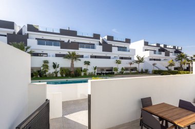 Manzaralı bahçeler, palmiye ağaçları ve berrak mavi gökyüzünün altında ortak bir yüzme havuzu olan modern bir apartman kompleksine bakan ahşap yemek masası olan özel bir teras..