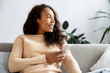 Kanepede oturan mutlu genç bir kadın, elinde akıllı telefon, başka tarafa bakıyor ve gülümsüyor. Evdeki iletişimin ve teknolojinin tadını çıkarıyor.