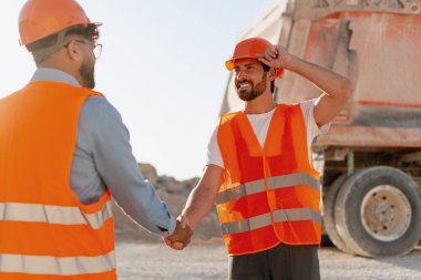 İnşaat işçileri taş ocağında turuncu şapka ve yelek giyip el sıkışıyorlar. Toplantı, takım çalışması konsepti
