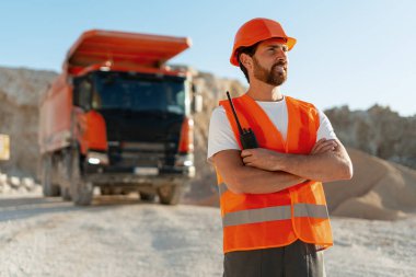 Turuncu güvenlik yeleği giymiş inşaat ustabaşı dışarıda kollarını çapraz yapıp endüstriyel alanda dikiliyor. Ağır makineler kavramı