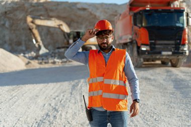 Turuncu miğfer ve yelek giyen kendinden emin Orta Doğulu mühendis madencilik alanında duruyor ve kameraya bakıyor. Taş ocağında profesyonel işçi. Ağır sanayi konsepti