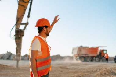 Turuncu güvenlik yeleği ve kasklı bir erkek inşaat denetçisi madencilik sahasında duruyor, ağır makine hareketini işaret etmek için elini kaldırıyor. Ağır sanayi konsepti