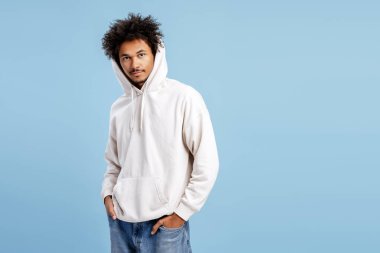 Yakışıklı, çekici Afro-Amerikan genç adam şık bir kapüşonlu giyiyor. Mavi arka planda izole edilmiş bir model. Alışveriş konsepti