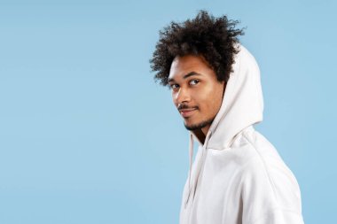 Yakışıklı, sakallı, kıvırcık saçlı, kafasına başlık takmış, kameraya bakan, mavi arka planda yalnız poz veren Amerikalı bir adam. Reklam kavramı