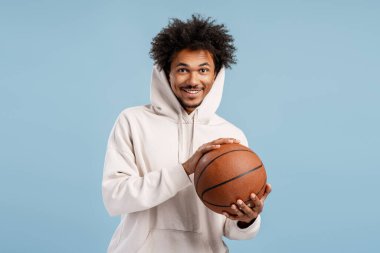 Basketbol topu tutan mutlu basketbolcu şık bir kapüşonlu giymiş kameraya bakıyor, mavi arka planda yalnız poz veriyor. Spor konsepti