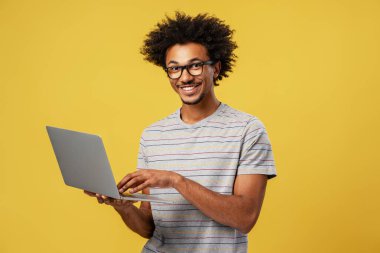 Gözlüklü mutlu bir serbest çalışan, klavyede yazı yazan, sarı arka planda izole edilmiş dizüstü bilgisayarı tutan. Bilgisayar üzerinde çalışan profesyonel bir işçi. Çevrimiçi teknoloji kavramı