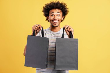 Heyecanlı, otantik Afro-Amerikan bir adam elinde kesekâğıdıyla sarı arka planda izole bir şekilde duran kameraya bakıyor. Alışveriş konsepti
