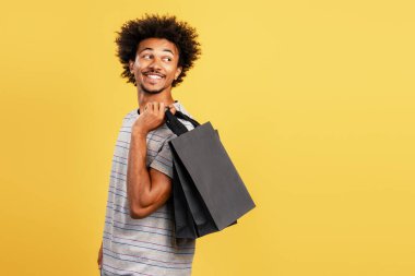 Mutlu, otantik Afro-Amerikan adam elinde siyah kese kağıdıyla sarı arka planda izole bir şekilde başka yere bakıyor. Alışveriş konsepti