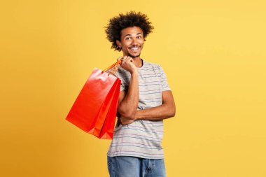 Kırmızı alışveriş çantası tutan yakışıklı Latin kıvırcık adam sarı arka planda izole edilmiş kameraya bakıyor. Alışveriş konsepti