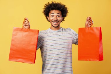Heyecanlı gülümseyen Afro-Amerikan adam elinde kırmızı kağıt torbalarla sarı arka planda izole edilmiş kameraya bakıyor. Alışveriş konsepti