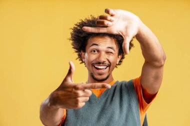 Gülümseyen mutlu Afro-Amerikalı adamın portresi. Sarı arka planda izole edilmiş kameraya parmaklarıyla bakıyor. Reklam kavramı