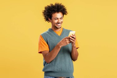 Sarı arka planda izole edilmiş mobil uygulama kullanarak cep telefonu tutan yakışıklı kıvırcık sakallı Afrikalı Amerikalı adamın portresi. Çevrimiçi teknoloji kavramı