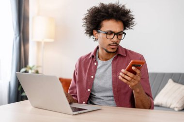 Ciddi, düşünceli, gözlüklü, cep telefonlu, mobil uygulama kullanan, evde dizüstü bilgisayarla çalışan Afro-Amerikan bir adam. Freelancer mesaj atmış. Çevrimiçi teknoloji kavramı