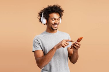 Gülümseyen Afrikalı Amerikalı adam kulaklıkla müzik dinliyor, cep telefonu kullanıyor, bej arka planda izole edilmiş mobil uygulama kullanıyor. Çevrimiçi teknoloji kavramı