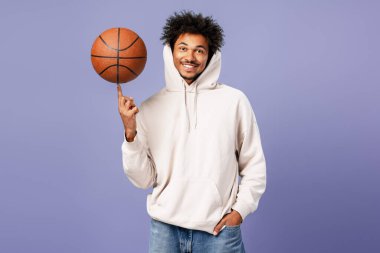 Basketbol topu tutan mutlu basketbolcu şık bir kapüşonlu giymiş kameraya bakıyor, mor arka planda yalnız poz veriyor. Spor konsepti