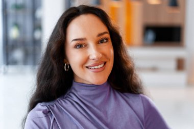 Gülümseyen genç bir kadın doğrudan kameraya bakıyor, içeride poz veriyor. Mutlu bir esmer kadının portresi.