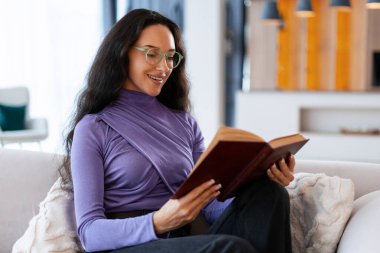 Kanepede oturan, kitap okuyan, gözlüklü ve mor gömlekli bir kadın, rahatlamanın tadını çıkarıyor.