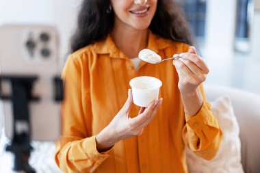 Genç bir kadın kendini akıllı bir telefonla çekerken yoğurt yiyor ve sağlıklı bir yaşam tarzı paylaşıyor.