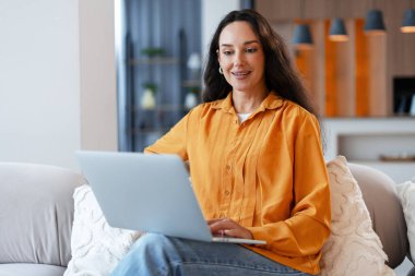 Kadın evde çalışırken dizüstü bilgisayara odaklanıyor, gülümsüyor. Serbest çalışan internetten iletişim kuruyor, koltukta rahatça oturuyor.