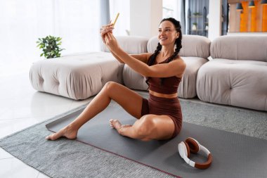 Spor kıyafetli bir kadın gülümsüyor, yoga minderinde selfie çekiyor, sosyal medyada spor programını paylaşıyor.