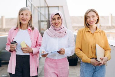 Profesyonel kadınlar çeşitliliği ve takım çalışmasını kutluyor, bir binanın çatısında yan yana duruyor, iş fikirlerini tartışıyor ve gülümsüyor.