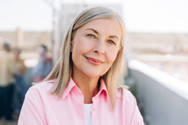 Gri saçlı yaşlı kadın gülümsüyor, kameraya bakıyor. Pembe gömlekli kendine güvenen olgun bir kadın dışarıda poz veriyor ve pozitif yaşlanma gösteriyor.