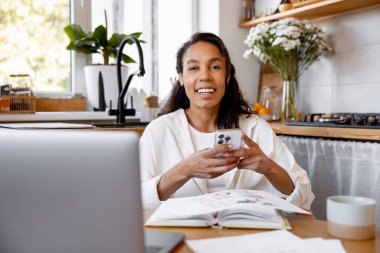 Mutfaktan uzaktan çalışan mutlu genç bir kadın akıllı telefon ve dizüstü bilgisayar kullanarak kameraya gülümsüyor.