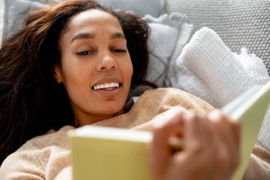 Genç siyahi kadın kanepeye uzanıp kitap okuyor. Boş zamanların ve içeride öğrenmenin tadını çıkarıyorum.