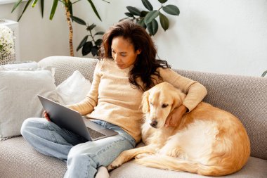 Golden Retriever köpeğiyle kanepede dizüstü bilgisayar kullanan bir kadın. Evden işe giderken refakatçiyle rahatlamak