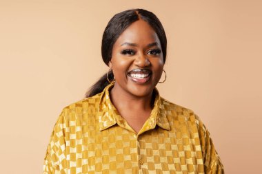 Siyah saçlı siyah kadın geri çekiliyor, halka küpeler takıyor ve altın bir gömlek giyiyor, kameraya sıcak bir şekilde gülümsüyor.