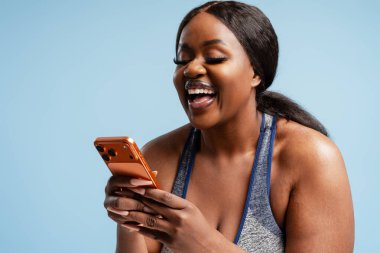 Spor kıyafetleri olan siyahi bir kadın açık mavi bir arka planda akıllı bir telefonla konuşurken yürekten gülüyor.