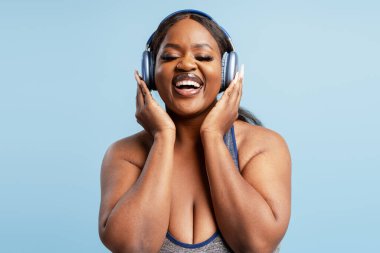 Kulaklık takan, açık mavi arka planda müzikle bağlantı kuran mutlu, genç siyahi kadın olumlu duygular hissediyor.