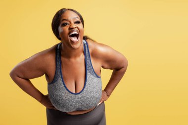 Spor giyimli neşeli, iri yarı siyahi bir kadın geniş bir alanda gülüyor, vücut pozitifliğini ve kendini kabulünü sarı arka planda kutluyor.