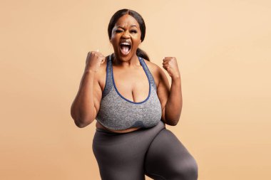 Spor kıyafetleri içindeki coşkulu siyahi kadın kaslarını esnetiyor, antrenmandan sonra başarıyı ve vücut pozitifliğini kutluyor.